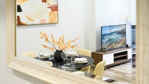 A dining area with a round table set for four, a vase with flowers, and a television displaying a coastal scene. - Modern 3-Bedroom Apartment Near Airport & Train (Mascot)