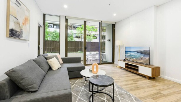 A modern living room with a grey sofa, a small round table, and a TV on a wooden cabinet. - Modern 3-Bedroom Apartment Near Airport & Train (Mascot)