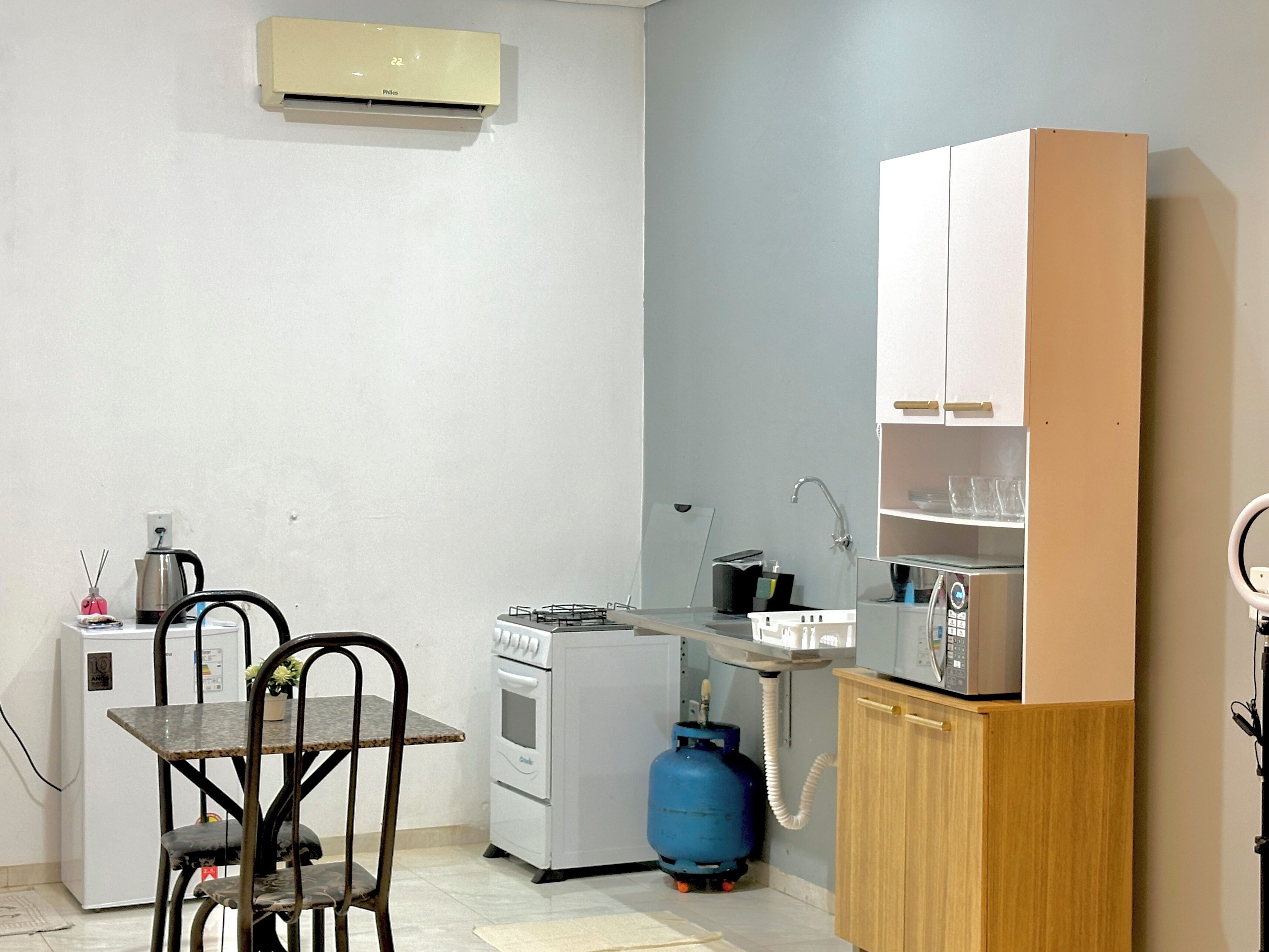 Une cuisine compacte équipée d’une cuisinière à gaz, d’un four et d’une bouteille de gaz bleue. On y trouve une petite table avec deux chaises, un réfrigérateur blanc et un meuble en bois avec des étagères.