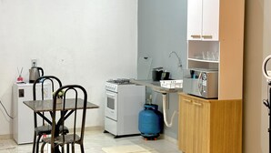 Cocina compacta con estufa a gas, horno y un cilindro de gas azul. Hay una mesa pequeña con dos sillas, un refrigerador blanco y un armario de madera con estantes.