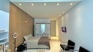 Een moderne slaapkamer met een bed, nachtkastje, lamp en een aan de muur bevestigde televisie.