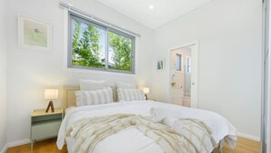 A bedroom with a large bed, a nightstand, a window with a view of greenery, and a door leading to another room. - Modern Riverwood Home with Parking & Easy Access (Riverwood)