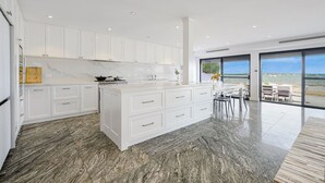 Una cocina moderna con armarios blancos y una isla grande, un comedor con vista al mar, y una sala de estar espaciosa con un ventanal.