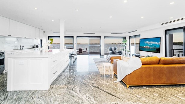 Cocina moderna con armarios blancos, encimera de mármol y una isla grande. Hay un comedor con sofá y televisor que muestra una vista panorámica.