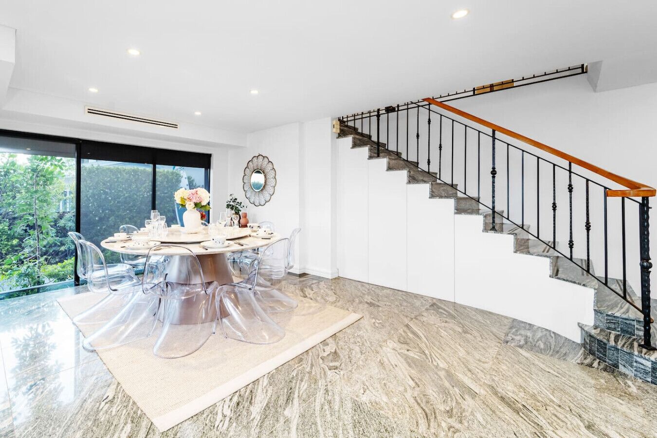 A modern dining area with a round table, clear chairs, and a view of greenery through large windows.