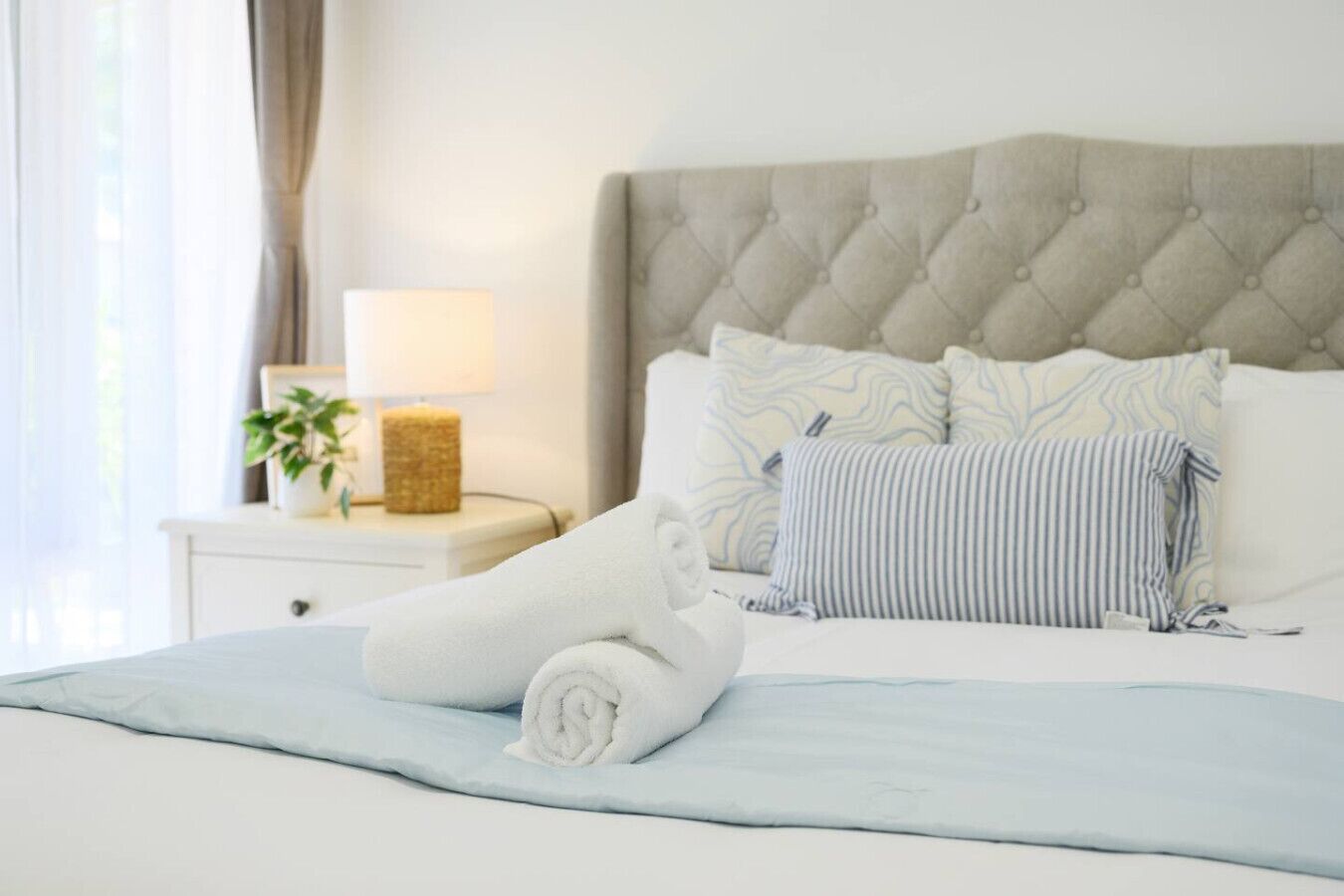 A neatly made bed with a rolled white towel, a grey tufted headboard, and striped pillows.