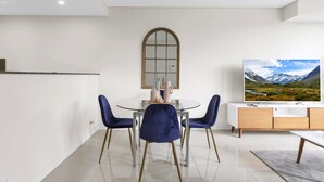 Uma área de jantar moderna com uma mesa redonda, quatro cadeiras azuis, uma mesa com tampo de vidro e um armário de madeira com uma TV em cima.