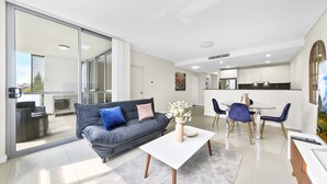 Uma sala de estar moderna com um sofá cinzento, uma mesa de centro branca e uma área de jantar com mesa de vidro e cadeiras azuis.