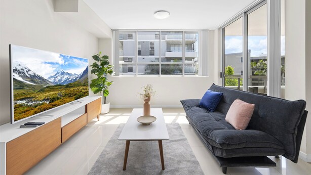 Uma sala de estar moderna com televisão de tela plana, sofá, mesa de centro e uma janela grande com vista para prédios e vegetação.