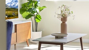 Quarto moderno com design minimalista, apresentando uma mesa branca, um armário de madeira, uma grande planta em vaso e um vaso com flores.