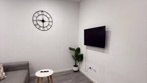 A modern living room with a grey sofa, a small round table, a wall-mounted clock, a potted plant, and a flat-screen TV. - Burwood Studio 1Br Near Train Station/Shopping (Burwood)