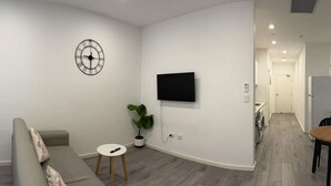 A modern living room with a grey sofa, a small white stool, a potted plant, and a wall-mounted TV. - Burwood Studio 1Br Near Train Station/Shopping (Burwood)