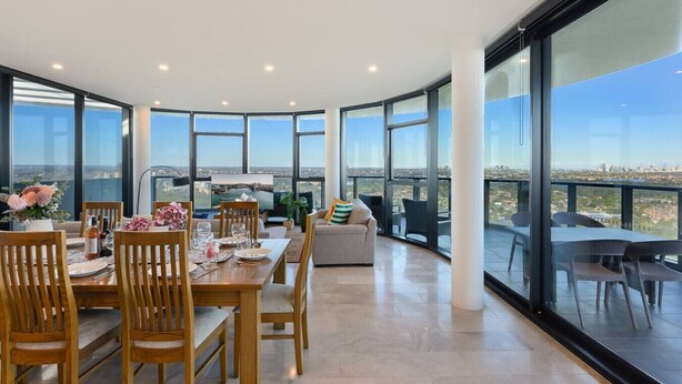 A spacious dining area with a high table and chairs, large windows offering a city view, and a balcony with outdoor seating.