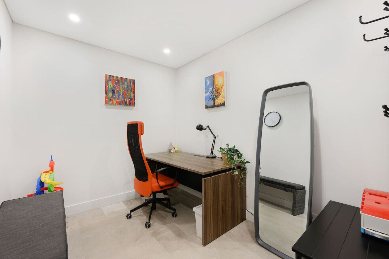 Un espace de bureau moderne avec un bureau en bois, une chaise de bureau orange, un grand miroir et deux tableaux abstraits accrochés au mur.