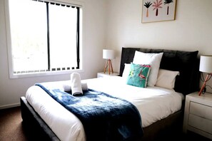 A neatly made bed with white linens, a blue throw blanket, and a white towel. There are two bedside lamps, a framed plant, and a window with blinds. - Kellyville Modern & Stylish House for family &work (Kellyville)