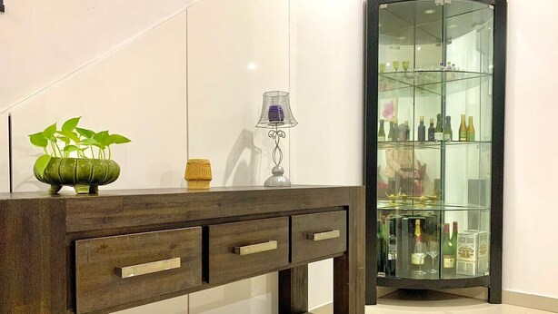Una consola de madera con tres cajones, una planta en maceta y una lámpara decorativa. Al fondo, un aparador de vidrio con varias botellas y vasos.