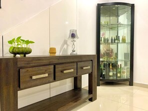 A wooden console table with three drawers, a potted plant, and a decorative lamp. In the background, there is a glass display cabinet with various bottles and glasses. - Near Train/Shopping at Bankstown (Bankstown)