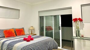 Un dormitorio con una cama, una mesita de noche, una lámpara, una puerta corrediza de vidrio y un florero con flores.