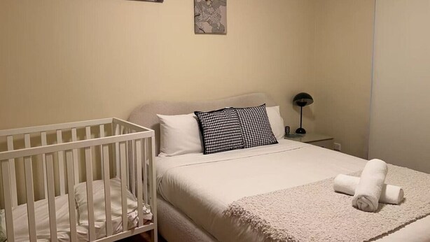 Une chambre avec un berceau, un lit recouvert d’une couette blanche, un oreiller noir et blanc, et deux œuvres d’art accrochées au mur.