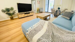 Ein modernes Wohnzimmer mit Flachbildfernseher, einem blauen Sofa, einem Couchtisch und einem großen Fenster mit Jalousien.