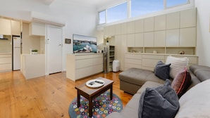 Ein modernes Wohnzimmer mit Sofa, Couchtisch und Fernseher. Es gibt eine Küchenzeile mit Schränken und einem Kühlschrank.