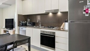 A modern kitchen with a gas stove, oven, and dishwasher. There is a dining table with chairs and a TV in the background. - 2BR APT near Train Station/4ppl/Couple/Business (Canterbury)