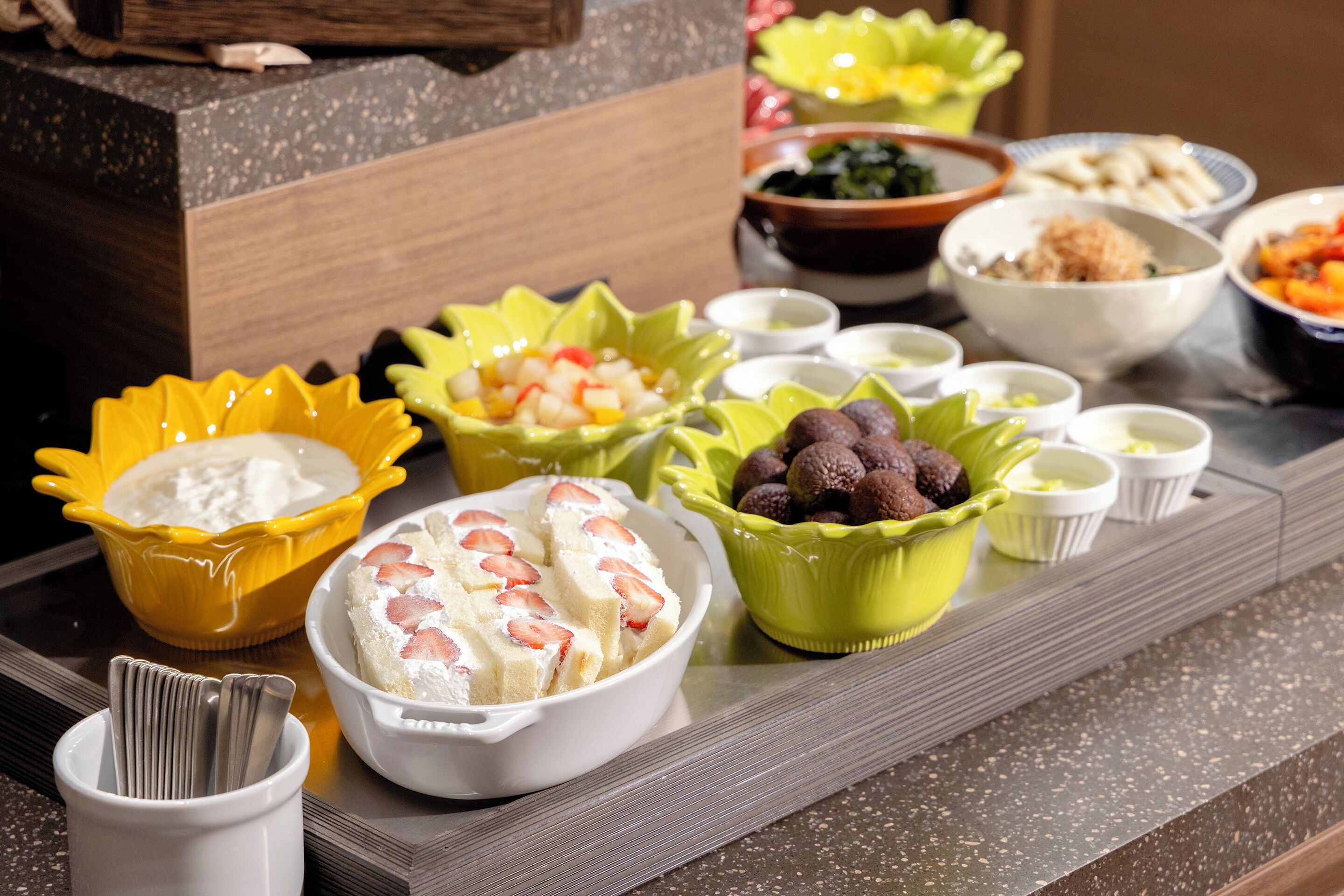 A table with various food items in bowls, spoons in a cup, and a wooden counter.