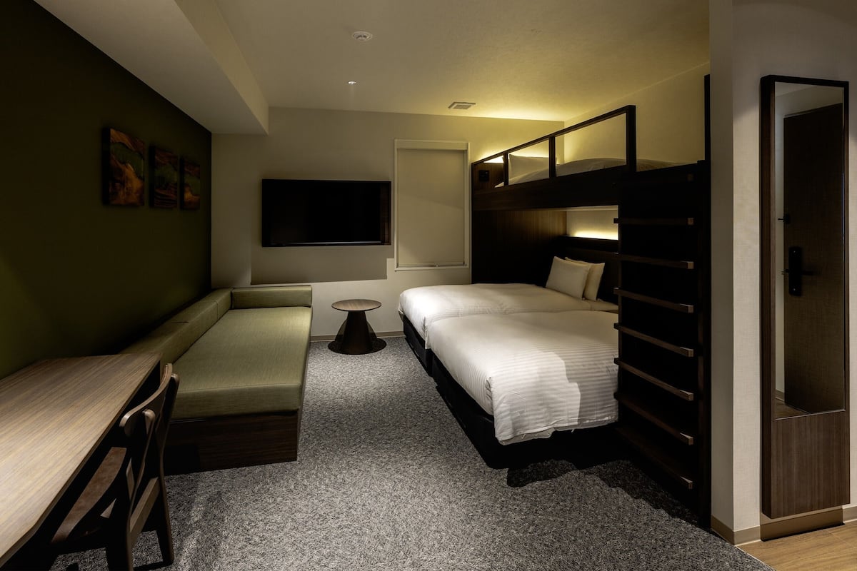 Habitación de hotel compacta con una cama litera, un escritorio, una silla, una pequeña mesa redonda y un televisor montado en la pared.