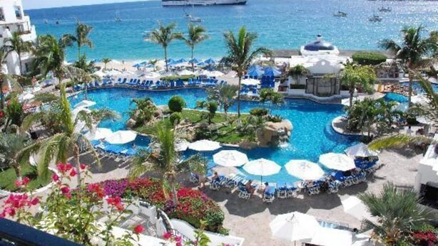 Un resort con piscina central rodeada de sillones de descanso y sombrillas, vista al océano y cruceros en la distancia.