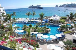 Un resort con piscina central rodeada de sillones de descanso y sombrillas, vista al océano y cruceros en la distancia.