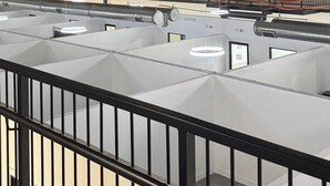Une grande chambre d’hôtel de style industriel, avec plusieurs lits disposés en rangées, un escalier central et des conduits apparents au plafond.