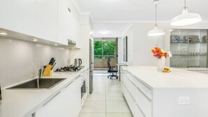 A modern kitchen with white cabinetry, a gas stove, and a coffee maker. There is a dining area with a chair and a vase of flowers. - Redfern 2Br Apt with balcony free parking near CBD (Redfern)