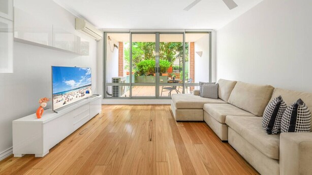 A modern living room with a flat-screen TV, a beige sectional sofa, and a glass door leading to an outdoor patio. - Redfern 2Br Apt with balcony free parking near CBD (Redfern)