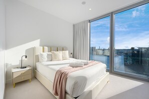 A modern bedroom with a large bed, a bedside table with a lamp, and a view of a cityscape through large windows. - Waterfront CBD luxury 3Br  with Parking for 8 ppl (Barangaroo)