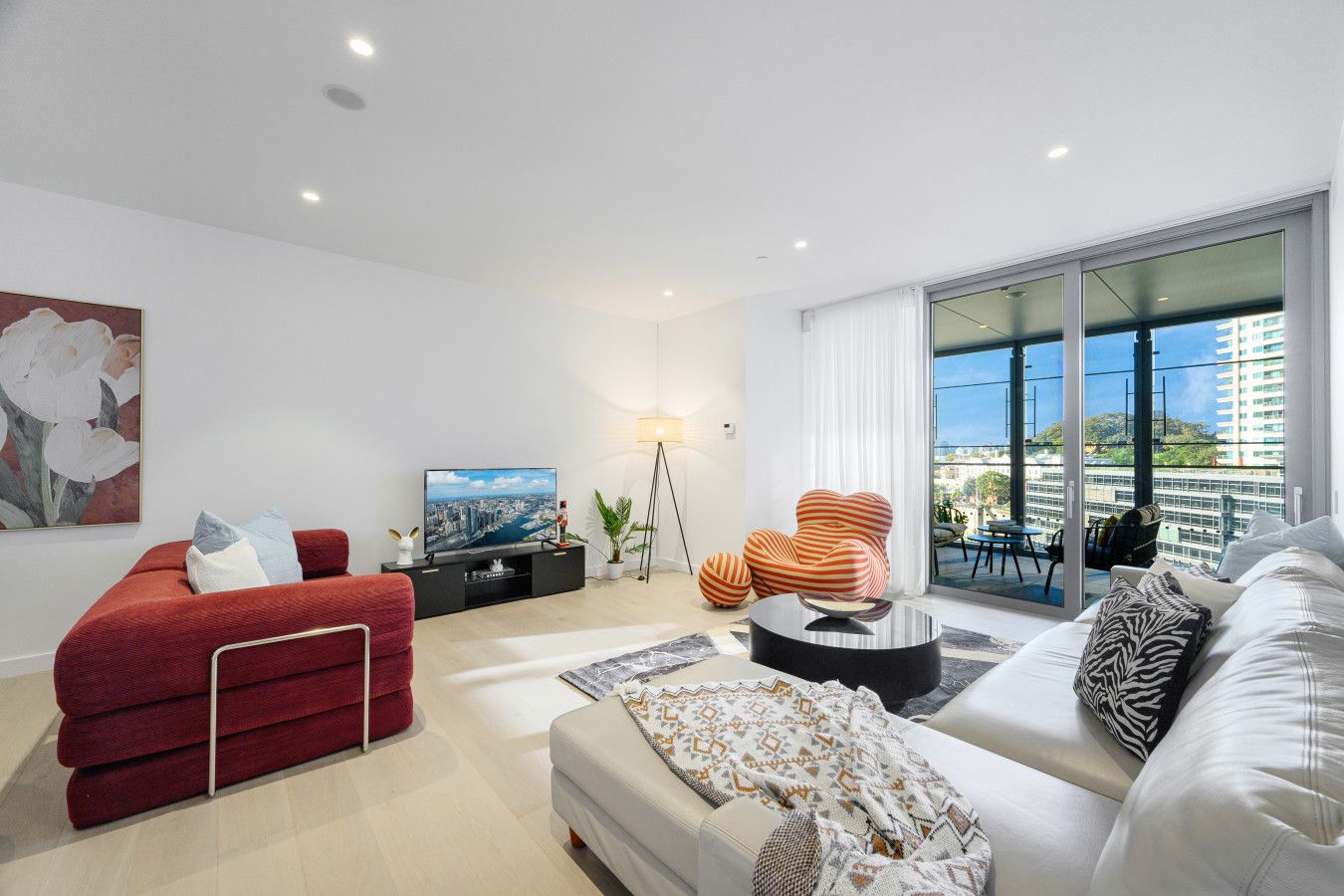 A modern living room with a flat-screen TV, red and white sofas, a coffee table, and a large window with a city view.