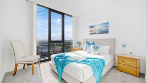 A modern hotel room with a large window, a bed with a blue throw, a chair, a nightstand, and a painting on the wall. - Parramatta 2br Apt Near Train Shopping Center (Parramatta)