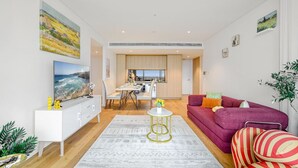 A modern living room with a sofa, a coffee table, and a TV. There is a dining area with a table and chairs in the background. - Parramatta 2br Apt Near Train Shopping Center (Parramatta)