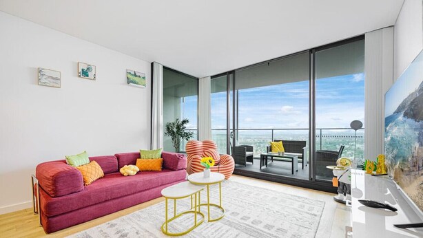 A modern living room with a red sofa, a small round table, and a large window offering a city view. - Parramatta 2br Apt Near Train Shopping Center (Parramatta)