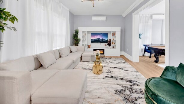 Un salon moderne avec un canapé d’angle beige, un fauteuil vert, un tapis à motifs de marbre, une table basse dorée et un téléviseur à écran plat fixé à un mur blanc.