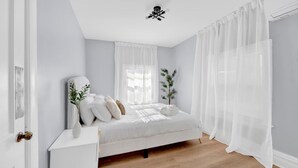 Une chambre à coucher comprenant un lit, une table de chevet, une fenêtre avec des rideaux légers et un ventilateur de plafond.
