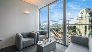 A modern hotel room with a large window offering a city view, grey upholstered seating, and a small side table with a book and a cup. - Waterfront CBD luxury 1Br with best view (Barangaroo)