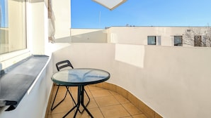 Ein kleiner Balkon mit einem Glastisch und Stühlen, einer weißen Wand und einem strahlend blauen Himmel.