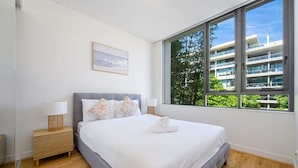 A bedroom with a large window, a bed with white bedding, a bedside table with a lamp, and a framed picture on the wall. - Rosebery near Airport & Train & Shops (Rosebery)