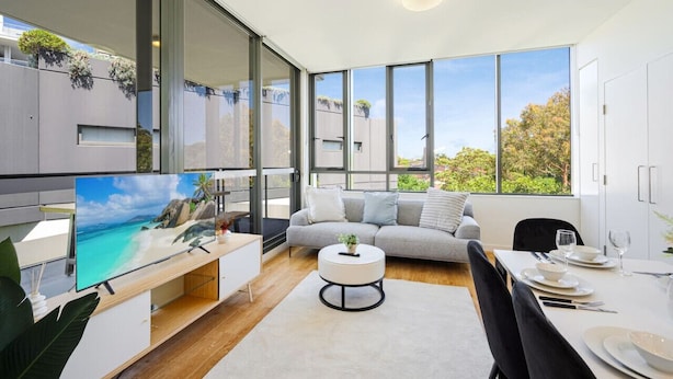 A modern living room with a large TV, a sofa, a dining table set for two, and large windows offering a view of greenery and buildings. - Rosebery near Airport & Train & Shops (Rosebery)