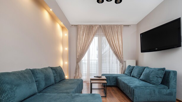 A modern living room with two teal sofas, a wooden floor, a flat-screen TV, and a window with sheer curtains. - Częstochowska Urban Comfort Apartment (Łódź)