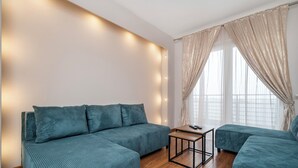 A modern living room with a sectional sofa, a small wooden table, and a large window with sheer curtains. - Częstochowska Urban Comfort Apartment (Łódź)