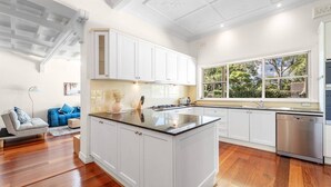 A modern kitchen with white cabinets, a central island, and a large window offering a view of greenery. - Stathfield Stylish 6br house with Pool/Tennis (Strathfield)