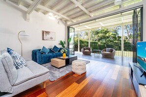 Un salon moderne avec un canapé, un fauteuil et une table basse. Une télévision se trouve sur le côté droit, et une grande fenêtre offre une vue sur l’extérieur.