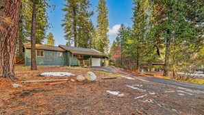 A cabin with a garage, surrounded by tall pine trees and a gravel driveway. - Evergreen Escape- Great location near downtown w/trailer parking and fully fenced yard (McCall)