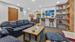 A living room with a leather sofa, a coffee table, and a bookshelf. There is a kitchen area in the background with blue cabinets and a bar stool. - Evergreen Escape- Great location near downtown w/trailer parking and fully fenced yard (McCall)